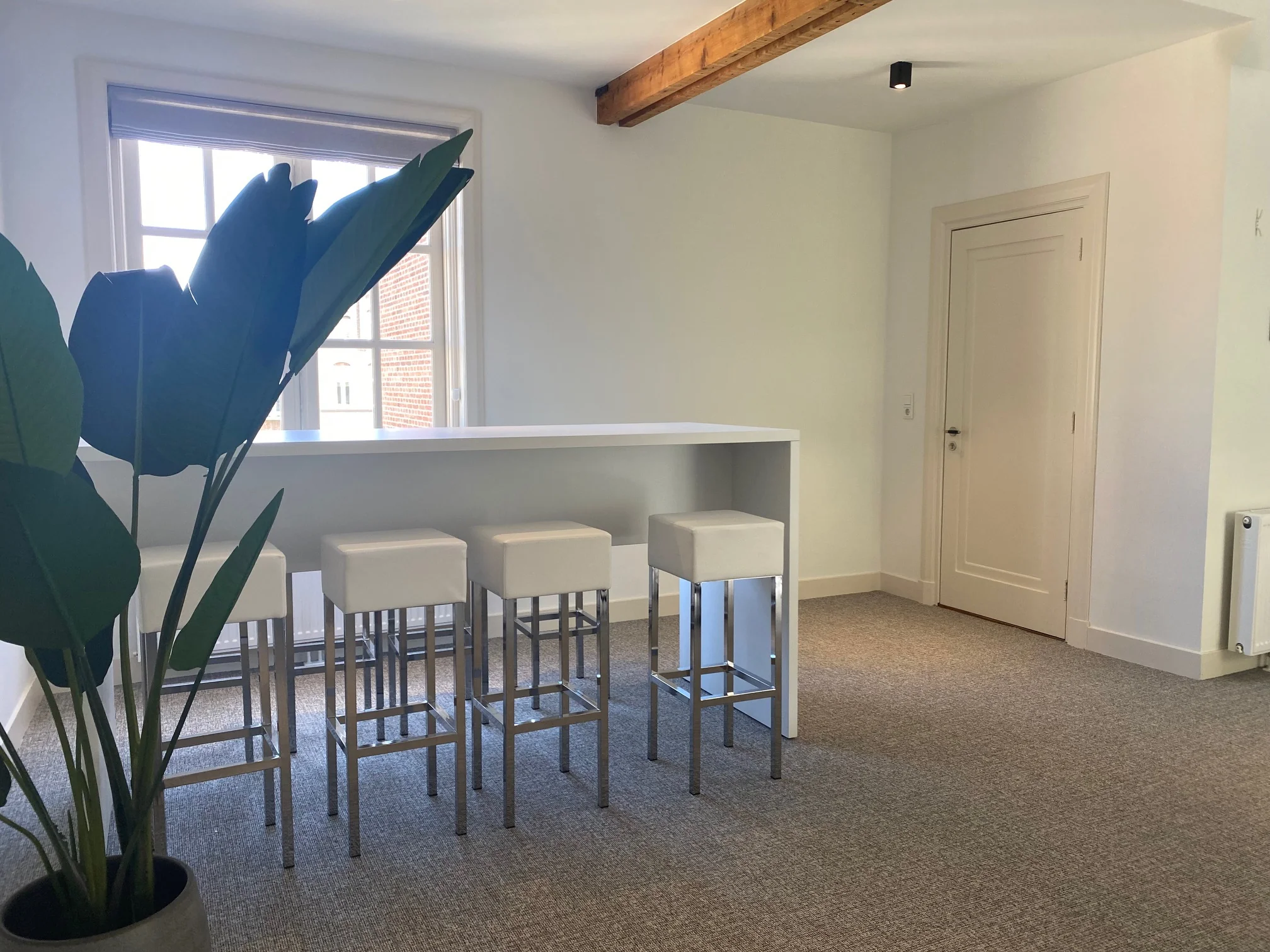 Moderne, lichte ruimte aan de Kleine Houtweg met hoge witte tafel, vier barkrukken en een grote kamerplant.